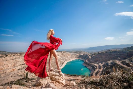 Side view of a beautiful sensual woman in a red long dress posing on a rock high above the lake in the afternoon. Against the background of the blue sky and the lake in the form of a heart.