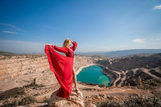 Side view of a beautiful sensual woman in a red long dress posing on a rock high above the lake in the afternoon. Against the background of the blue sky and the lake in the form of a heart.