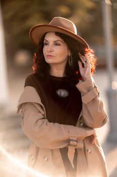 Outdoor fashion portrait of young elegant fashionable brunette woman, model in stylish hat, choker and light raincoat posing at sunset in European city