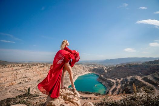 Side view of a beautiful sensual woman in a red long dress posing on a rock high above the lake in the afternoon. Against the background of the blue sky and the lake in the form of a heart.