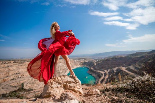 Side view of a beautiful sensual woman in a red long dress posing on a rock high above the lake in the afternoon. Against the background of the blue sky and the lake in the form of a heart.