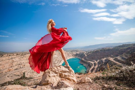 Side view of a beautiful sensual woman in a red long dress posing on a rock high above the lake in the afternoon. Against the background of the blue sky and the lake in the form of a heart.