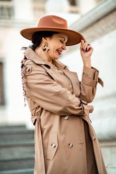 Outdoor fashion portrait of young elegant fashionable brunette woman, model in stylish hat, choker and light raincoat posing at sunset in European city