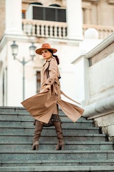 Outdoor fashion portrait of young elegant fashionable brunette woman, model in stylish hat, choker and light raincoat posing at sunset in European city