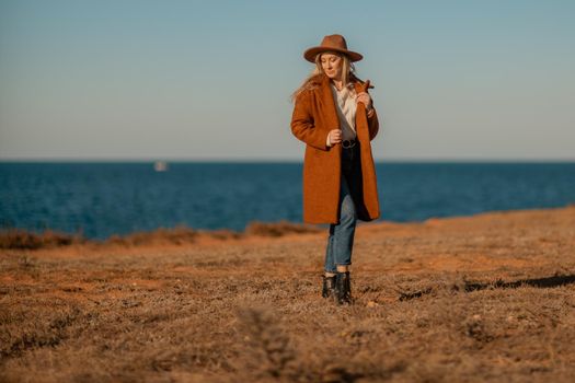 A woman walking along the coast near the sea. An elegant lady in a brown coat and a hat with fashionable makeup walks on the seashore.