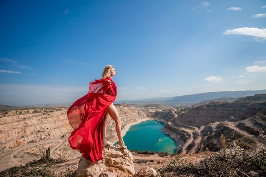 Side view of a beautiful sensual woman in a red long dress posing on a rock high above the lake in the afternoon. Against the background of the blue sky and the lake in the form of a heart.