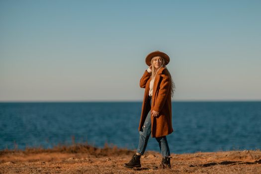 A woman walking along the coast near the sea. An elegant lady in a brown coat and a hat with fashionable makeup walks on the seashore.