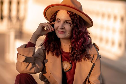 Outdoor fashion portrait of young elegant fashionable brunette woman, model in stylish hat, choker and light raincoat posing at sunset in European city