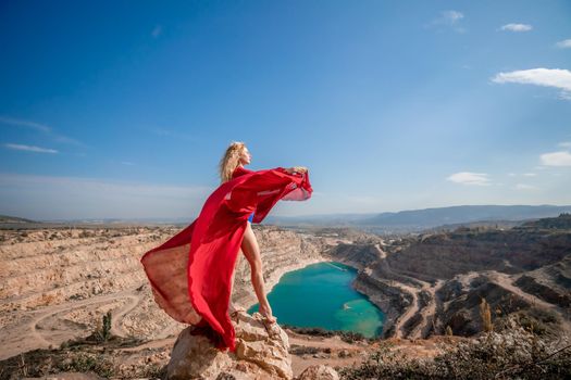 Side view of a beautiful sensual woman in a red long dress posing on a rock high above the lake in the afternoon. Against the background of the blue sky and the lake in the form of a heart.