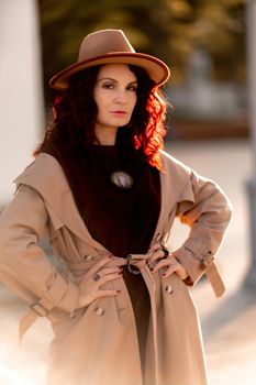 Outdoor fashion portrait of young elegant fashionable brunette woman, model in stylish hat, choker and light raincoat posing at sunset in European city