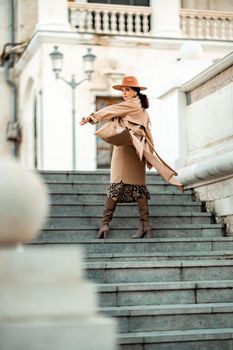 Outdoor fashion portrait of young elegant fashionable brunette woman, model in stylish hat, choker and light raincoat posing at sunset in European city