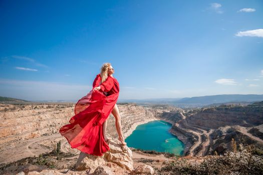 Side view of a beautiful sensual woman in a red long dress posing on a rock high above the lake in the afternoon. Against the background of the blue sky and the lake in the form of a heart.