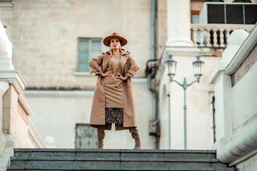 Outdoor fashion portrait of young elegant fashionable brunette woman, model in stylish hat, choker and light raincoat posing at sunset in European city