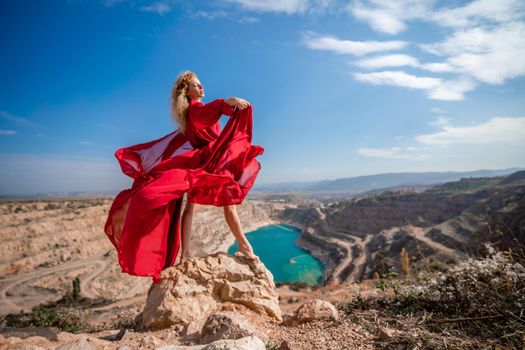 Side view of a beautiful sensual woman in a red long dress posing on a rock high above the lake in the afternoon. Against the background of the blue sky and the lake in the form of a heart.