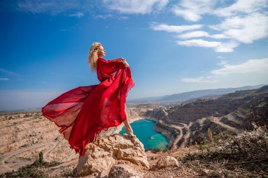 Side view of a beautiful sensual woman in a red long dress posing on a rock high above the lake in the afternoon. Against the background of the blue sky and the lake in the form of a heart.