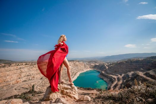 Side view of a beautiful sensual woman in a red long dress posing on a rock high above the lake in the afternoon. Against the background of the blue sky and the lake in the form of a heart.