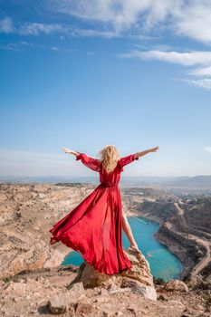 Rear view of a beautiful sensual woman in a red long dress posing on a rock high above the lake in the afternoon. Against the background of the blue sky and the lake in the shape of a heart.