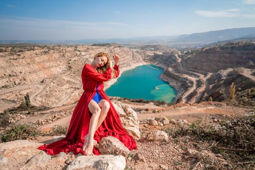 A beautiful girl in a red long dress, Sits on a rock high above the lake in the afternoon. Against the background of the blue sky and the lake in the shape of a heart