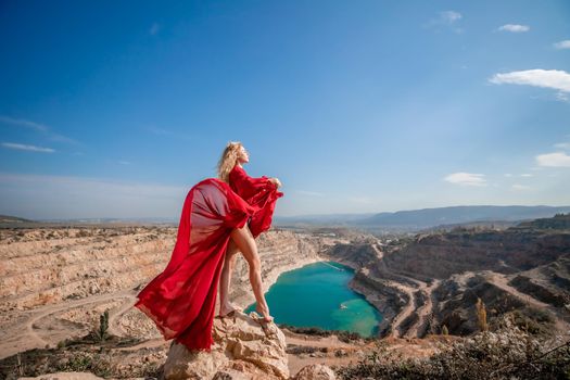 Side view of a beautiful sensual woman in a red long dress posing on a rock high above the lake in the afternoon. Against the background of the blue sky and the lake in the form of a heart.