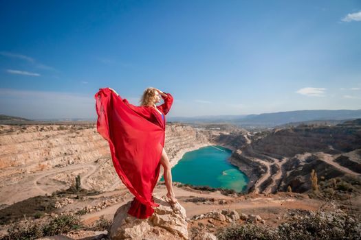 Side view of a beautiful sensual woman in a red long dress posing on a rock high above the lake in the afternoon. Against the background of the blue sky and the lake in the form of a heart.