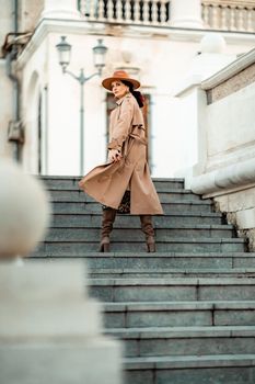 Outdoor fashion portrait of young elegant fashionable brunette woman, model in stylish hat, choker and light raincoat posing at sunset in European city