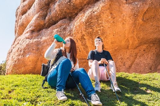 two women hikers, resting sitting on the grass. young women on vacation. woman drinking water from a canteen.travel person. woman walking in nature. background of a mountain with green plants illuminated by sun rays.