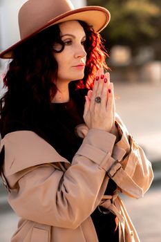 Outdoor fashion portrait of young elegant fashionable brunette woman, model in stylish hat, choker and light raincoat posing at sunset in European city