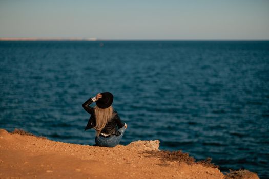 A serious blonde girl in a stylish black leather jacket and a black hat is sitting with her back to the seashore.