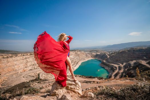 Side view of a beautiful sensual woman in a red long dress posing on a rock high above the lake in the afternoon. Against the background of the blue sky and the lake in the form of a heart.