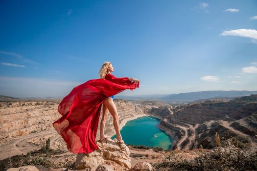 Side view of a beautiful sensual woman in a red long dress posing on a rock high above the lake in the afternoon. Against the background of the blue sky and the lake in the form of a heart.