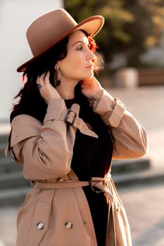 Outdoor fashion portrait of young elegant fashionable brunette woman, model in stylish hat, choker and light raincoat posing at sunset in European city