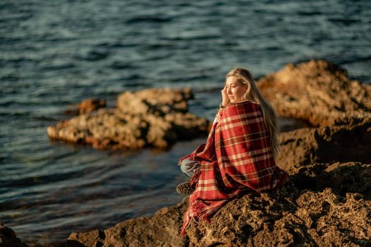 Attractive blonde Caucasian woman enjoying time on the beach at sunset, sitting in a blanket and looking to the side, with the sunset sky and sea in the background. Beach vacation