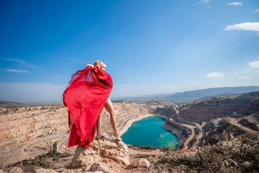 Side view of a beautiful sensual woman in a red long dress posing on a rock high above the lake in the afternoon. Against the background of the blue sky and the lake in the form of a heart.