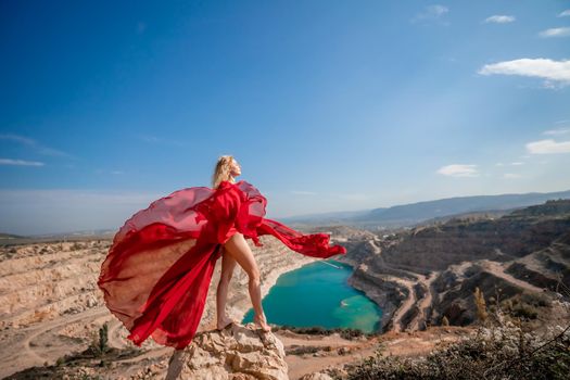 Side view of a beautiful sensual woman in a red long dress posing on a rock high above the lake in the afternoon. Against the background of the blue sky and the lake in the form of a heart.