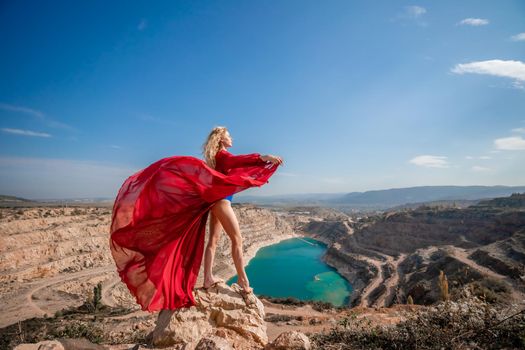 Side view of a beautiful sensual woman in a red long dress posing on a rock high above the lake in the afternoon. Against the background of the blue sky and the lake in the form of a heart.