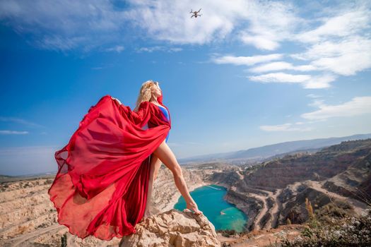 Side view of a beautiful sensual woman in a red long dress posing on a rock high above the lake in the afternoon. Against the background of the blue sky and the lake in the form of a heart.