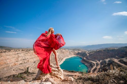 Side view of a beautiful sensual woman in a red long dress posing on a rock high above the lake in the afternoon. Against the background of the blue sky and the lake in the form of a heart.