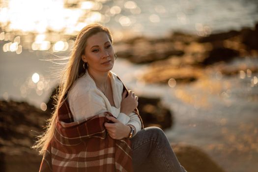 Attractive blonde Caucasian woman enjoying time on the beach at sunset, sitting in a blanket and looking to the side, with the sunset sky and sea in the background. Beach vacation