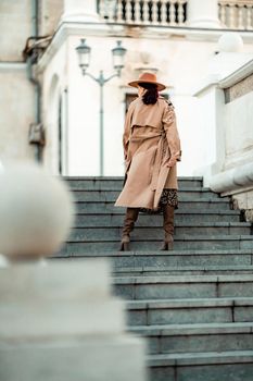 Outdoor fashion portrait of young elegant fashionable brunette woman, model in stylish hat, choker and light raincoat posing at sunset in European city