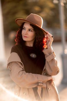 Outdoor fashion portrait of young elegant fashionable brunette woman, model in stylish hat, choker and light raincoat posing at sunset in European city