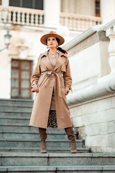 Outdoor fashion portrait of young elegant fashionable brunette woman, model in stylish hat, choker and light raincoat posing at sunset in European city