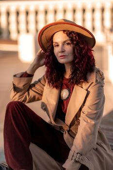 Outdoor fashion portrait of young elegant fashionable brunette woman, model in stylish hat, choker and light raincoat posing at sunset in European city