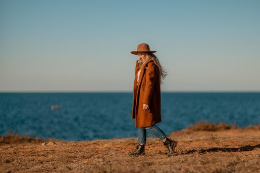 A woman walking along the coast near the sea. An elegant lady in a brown coat and a hat with fashionable makeup walks on the seashore.