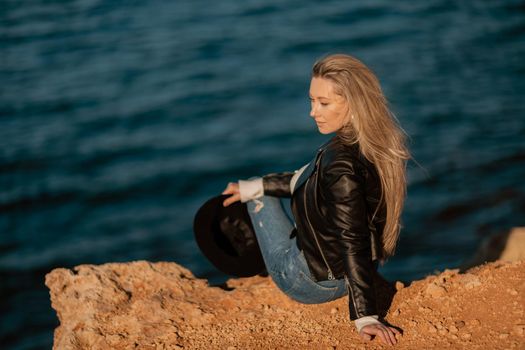 A blonde girl in a stylish black leather jacket is sitting with her back to the seashore