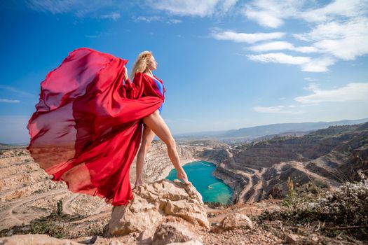 Side view of a beautiful sensual woman in a red long dress posing on a rock high above the lake in the afternoon. Against the background of the blue sky and the lake in the form of a heart.