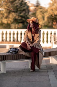 Outdoor fashion portrait of young elegant fashionable brunette woman, model in stylish hat, choker and light raincoat posing at sunset in European city