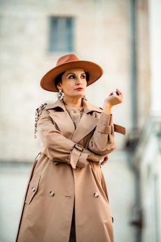 Outdoor fashion portrait of young elegant fashionable brunette woman, model in stylish hat, choker and light raincoat posing at sunset in European city