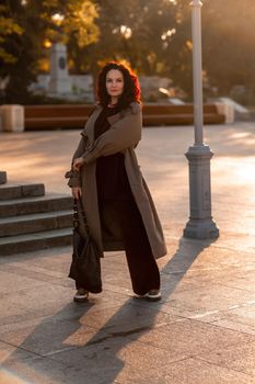 Outdoor fashion portrait of young elegant fashionable brunette woman, model in stylish hat, choker and light raincoat posing at sunset in European city