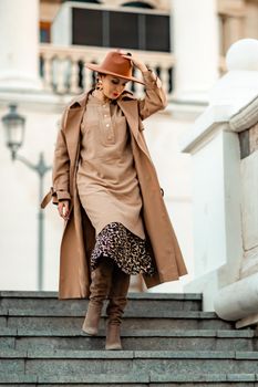 Outdoor fashion portrait of young elegant fashionable brunette woman, model in stylish hat, choker and light raincoat posing at sunset in European city