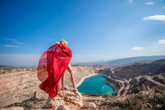 Side view of a beautiful sensual woman in a red long dress posing on a rock high above the lake in the afternoon. Against the background of the blue sky and the lake in the form of a heart.